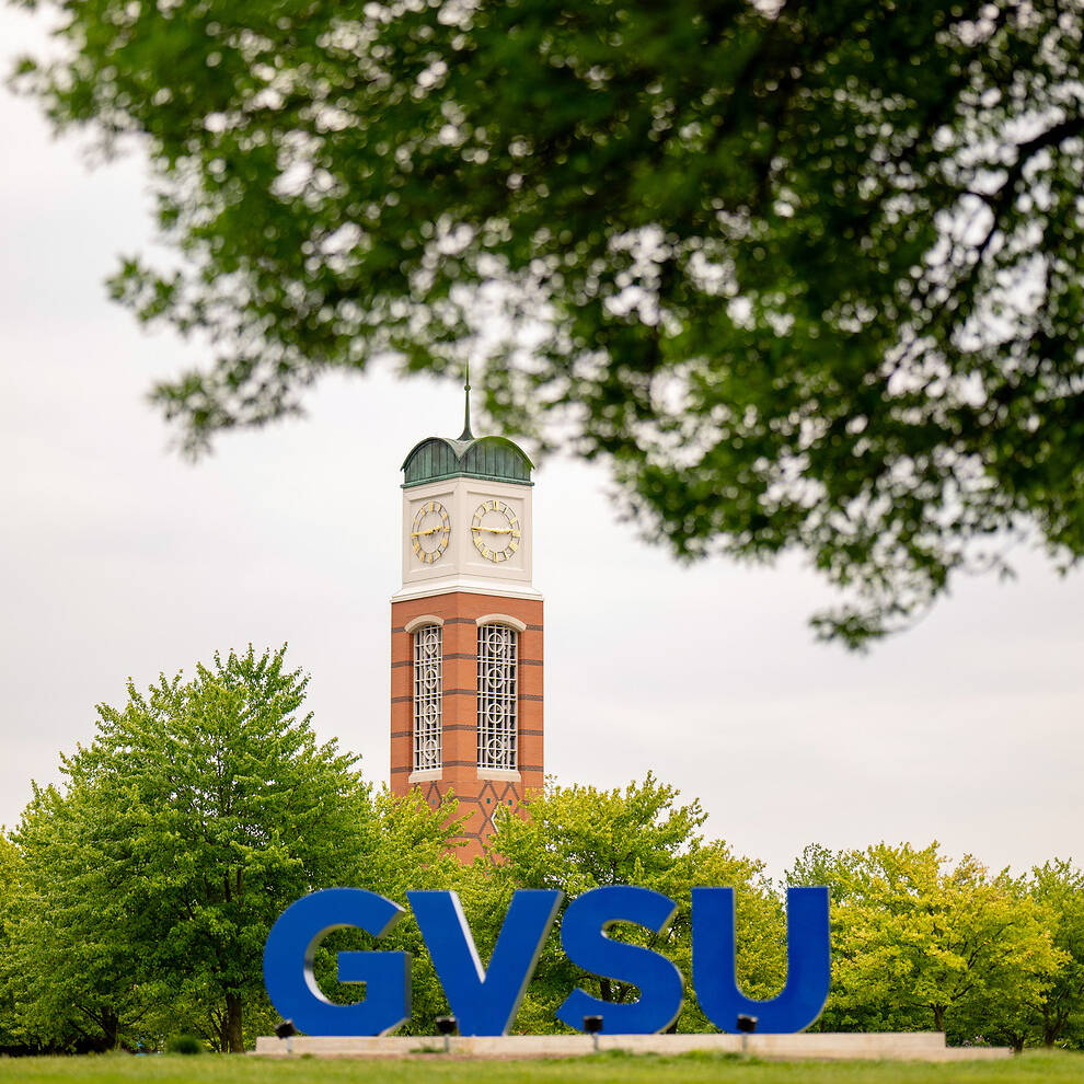 A clock tower stands behind large blue "GVSU" letters, framed by green trees and a grassy field.
